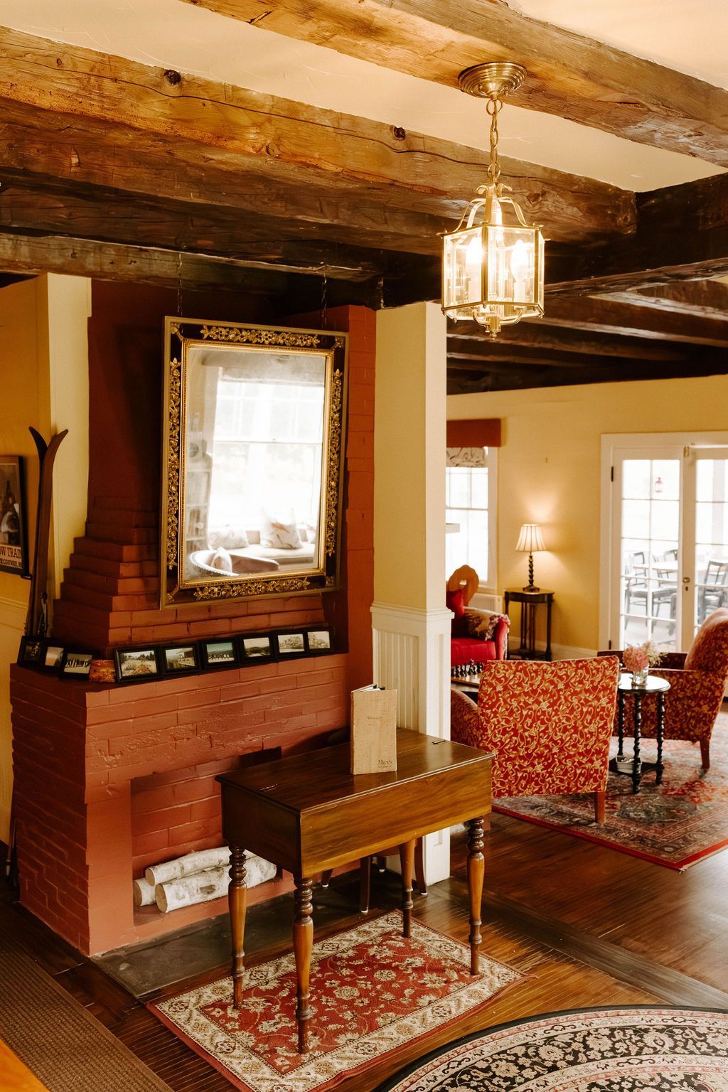 Cozy living room with exposed beams, brick fireplace, large mirror, and patterned armchairs by a window.