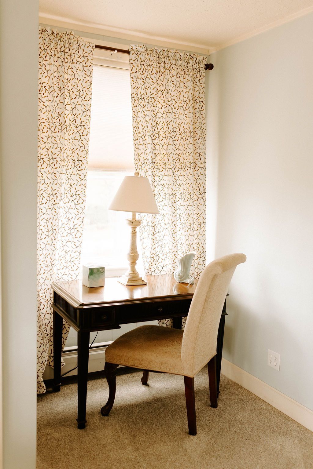 Small home office with desk, lamp, chair, and patterned curtains by a bright window.