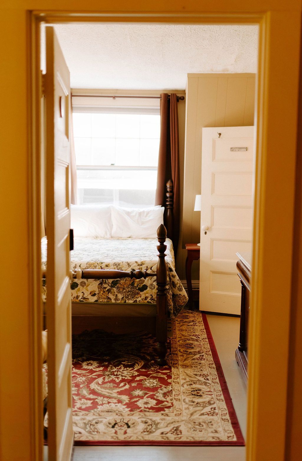 Narrow hallway with patterned rug leading to a bedroom with a bed, window, and white dresser