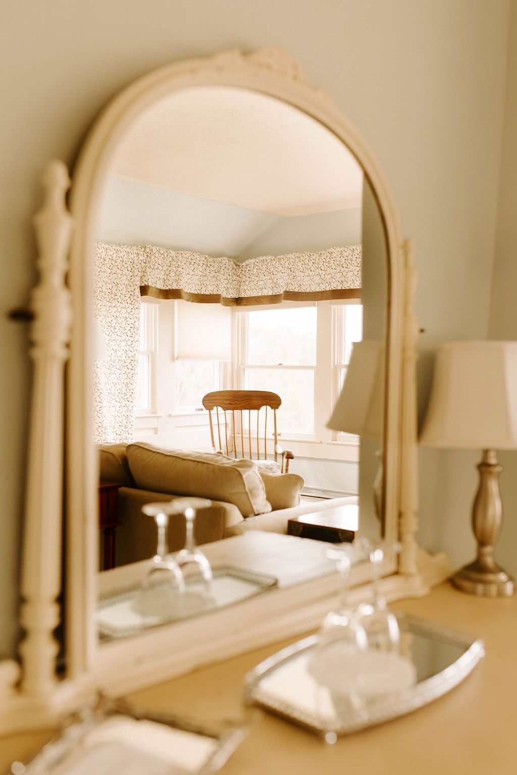 Bright bedroom reflected in an arched mirror on a dresser, with a lamp and tray in the foreground