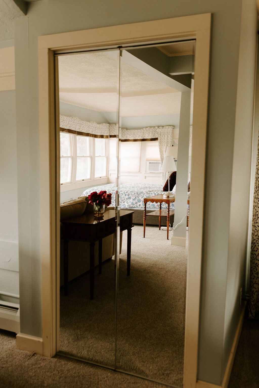 Mirror on a hallway wall reflecting a bright room with windows, a table, and chairs