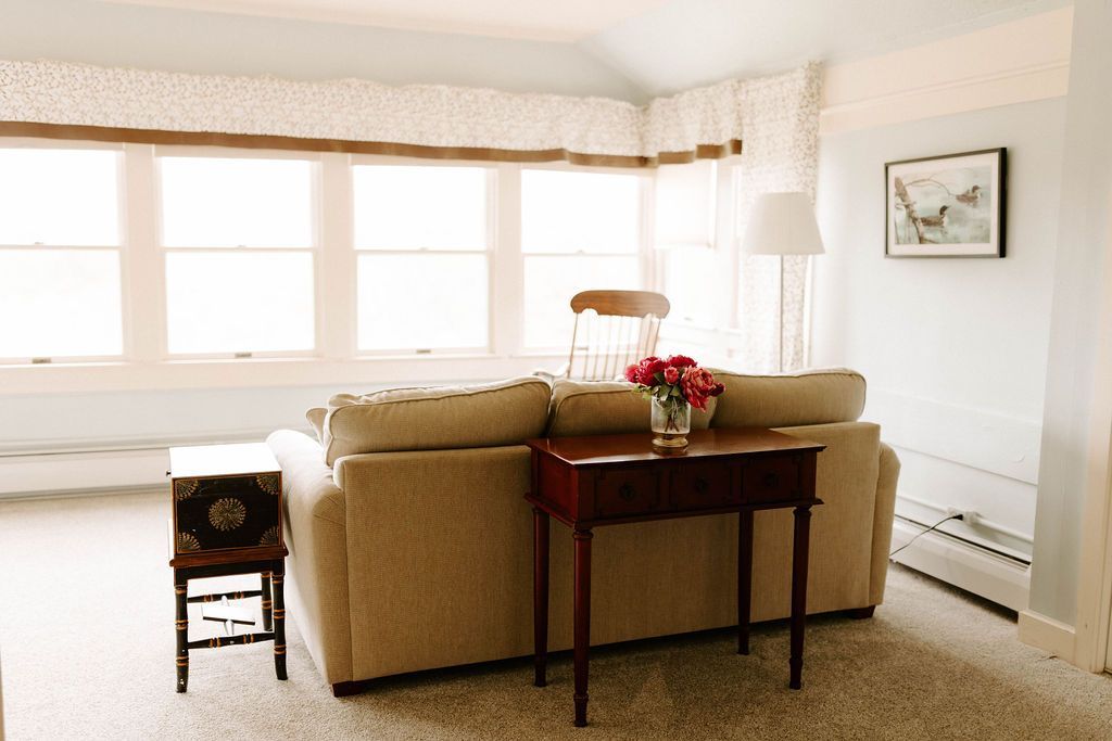 Bright living room with beige sofa, wooden table, flowers, large windows, and framed wall art.