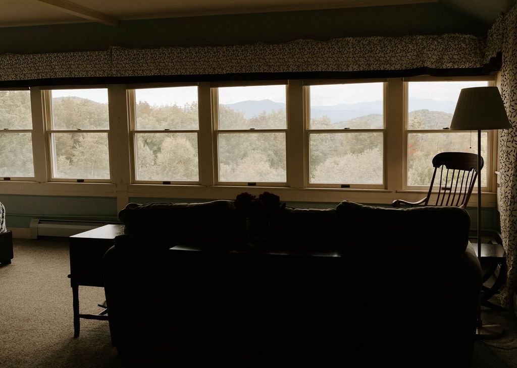 Dim living room with a large sectional sofa facing wide windows and a view of trees outside