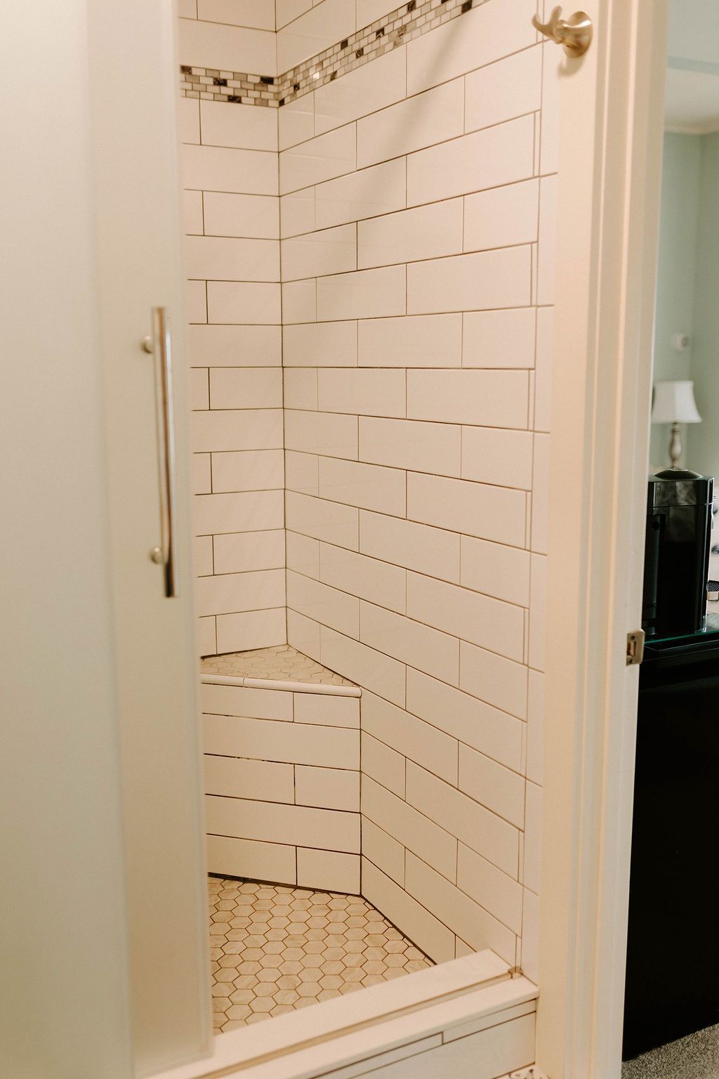 Small white tiled shower with glass door and brass handle, viewed from the bathroom doorway.