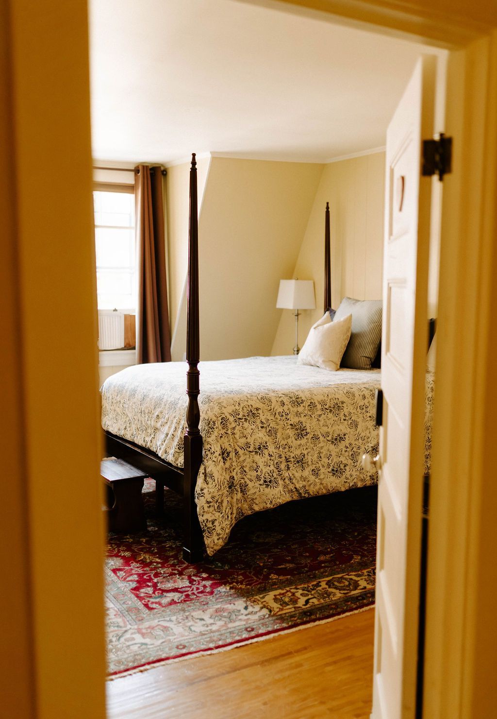 Cozy bedroom with a four-poster bed, patterned rug, and window light near an open doorway