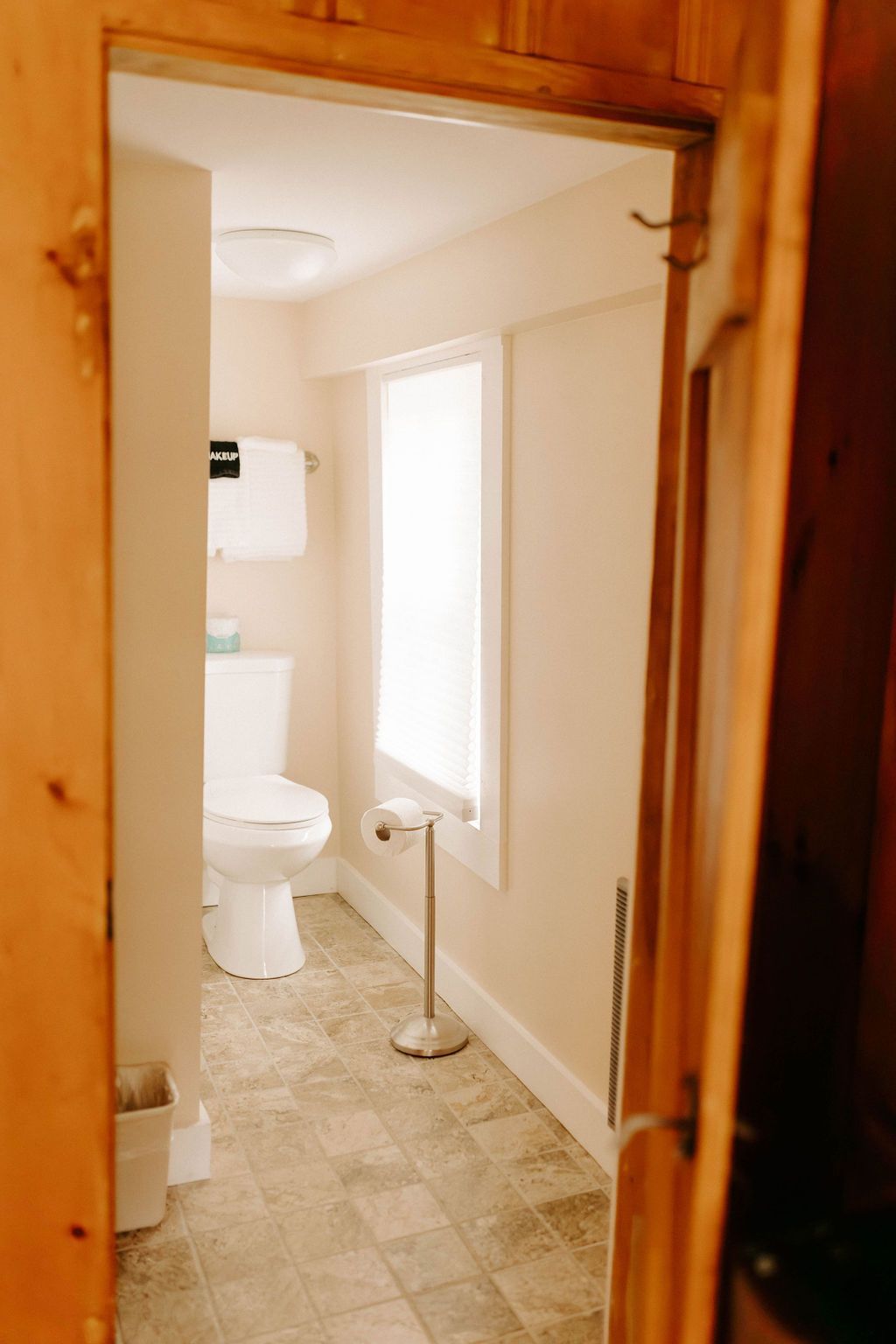 View through a wooden door into a small beige bathroom with a toilet, window, and tiled floor.