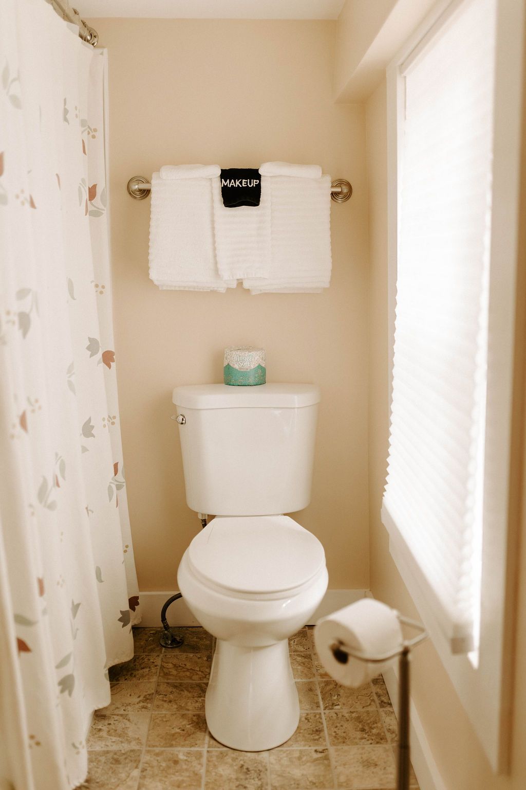 Small beige bathroom with a toilet, wall-mounted towel rack, and toilet paper holder near a window