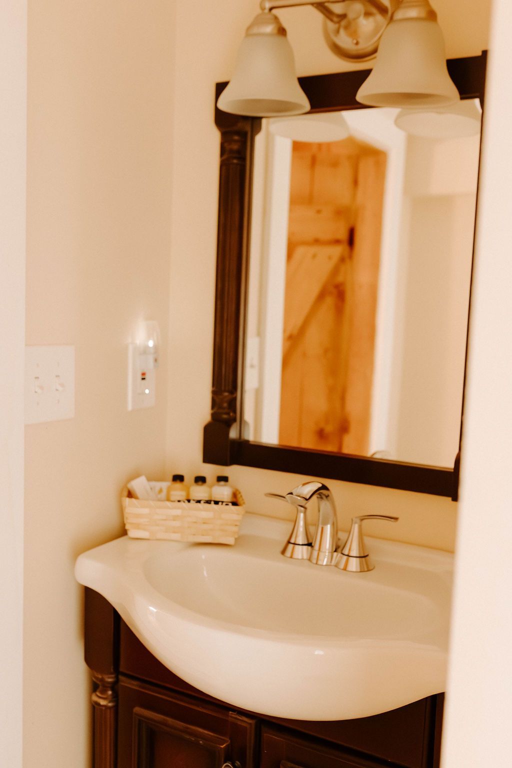 Small bathroom sink with dark wood mirror frame, cream walls, and two light fixtures above.