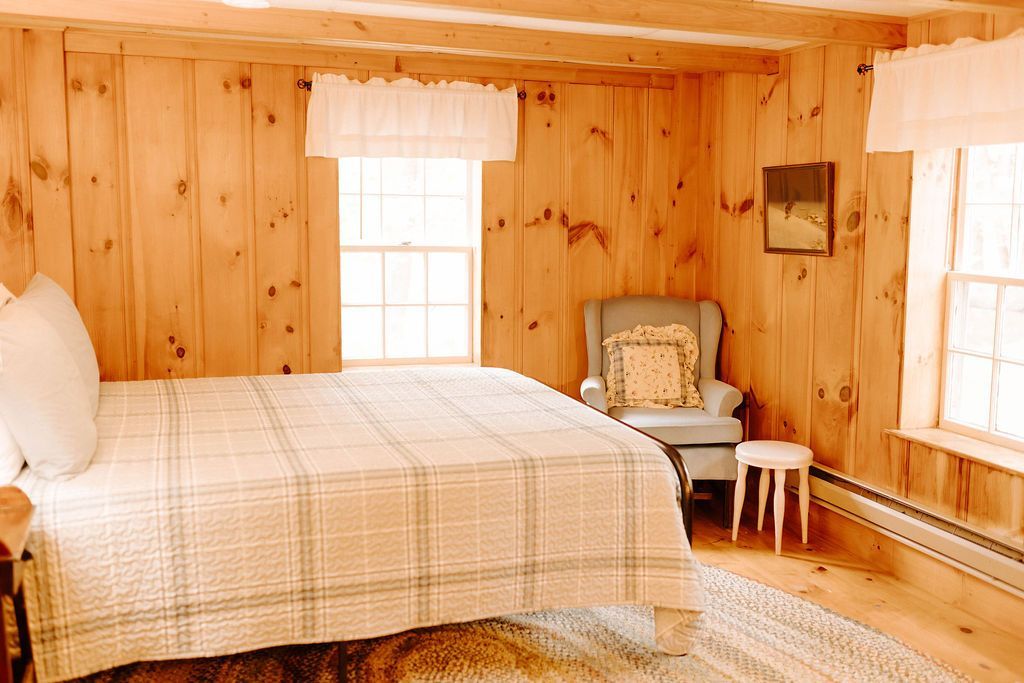 Cozy wood-paneled bedroom with a bed, chair, small table, and sunlight through large windows