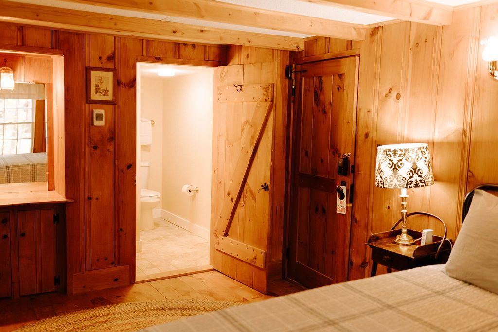 Rustic wood-paneled bedroom with open bathroom door, bed, and lit bedside lamp