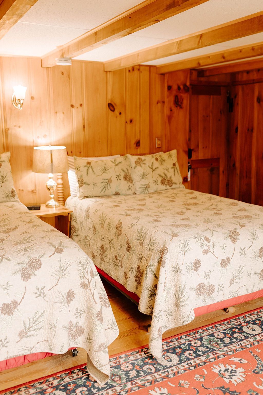 Cozy wood-paneled bedroom with two floral-covered beds, lamp, and patterned rug