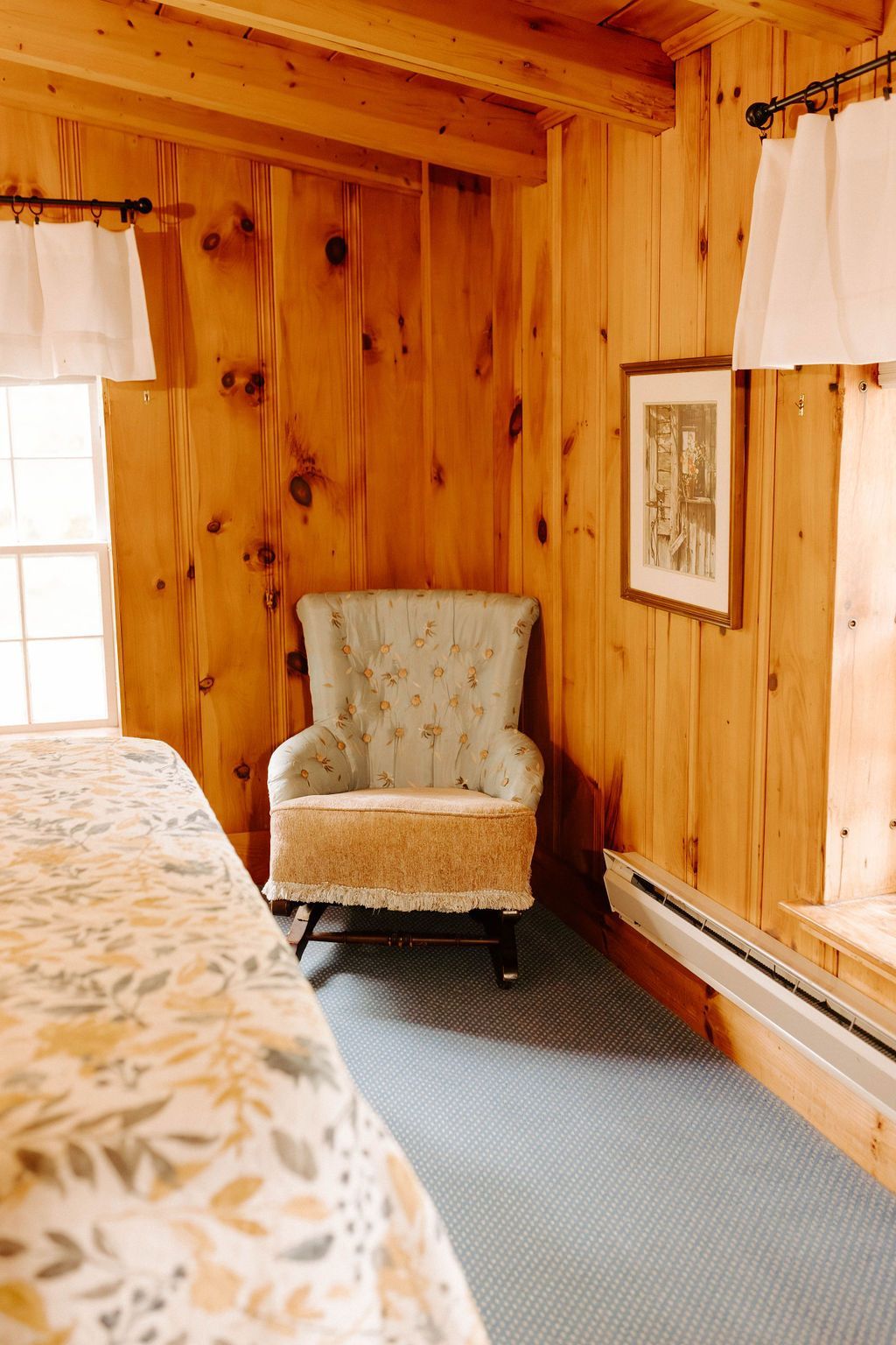Small wood-paneled bedroom with a plaid armchair, bed edge, two windows, and a framed picture