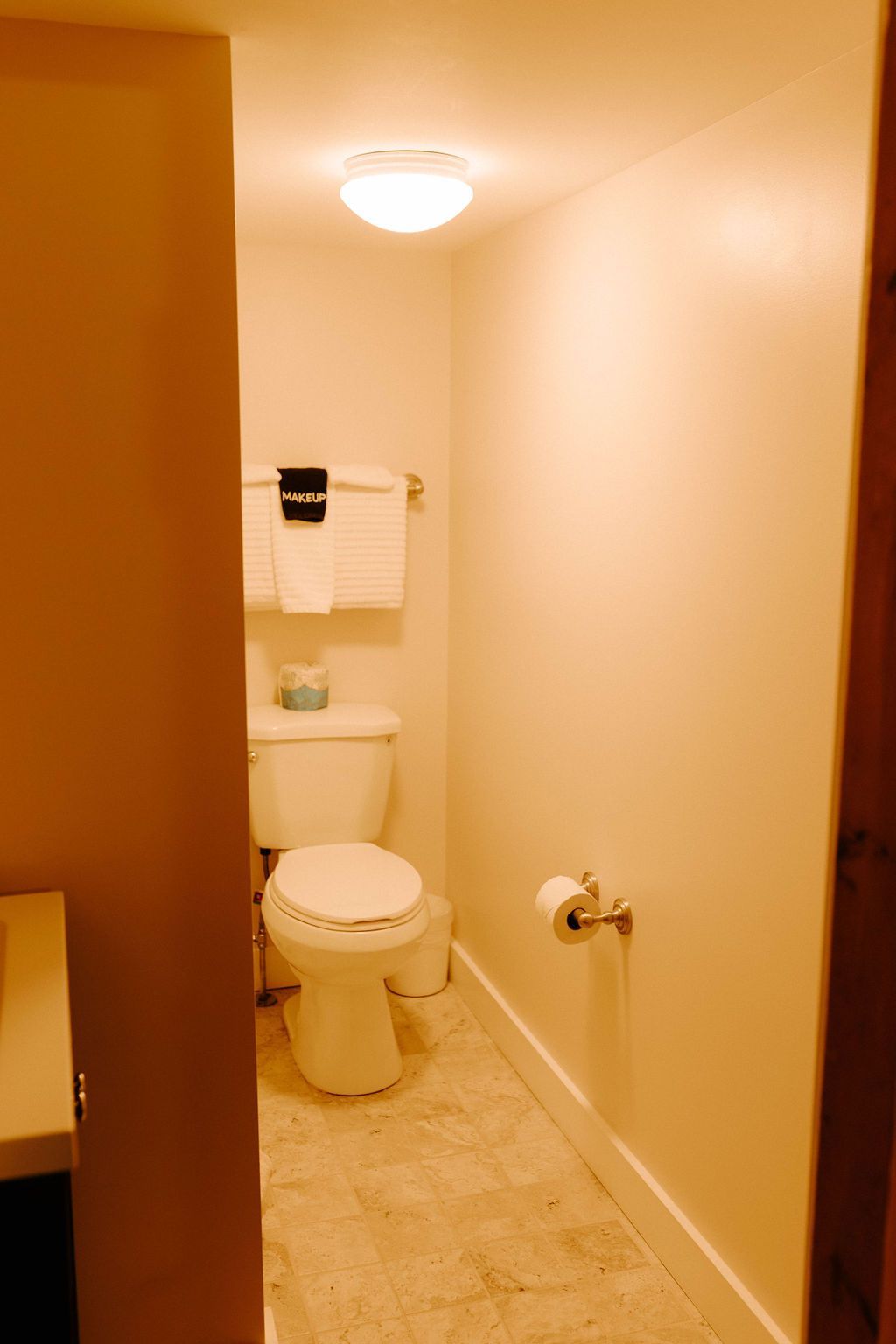 Small beige bathroom with toilet, sink, towel rack, and ceiling light.