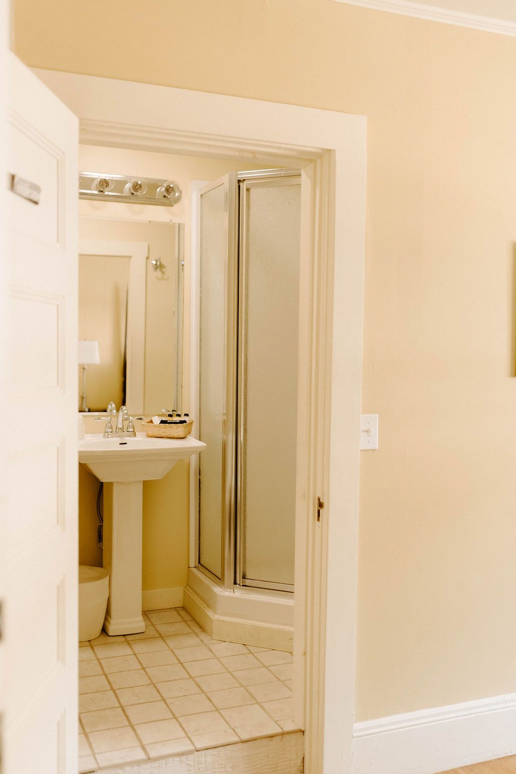 Bathroom hallway with beige walls, tiled floor, pedestal sink, and glass shower door.