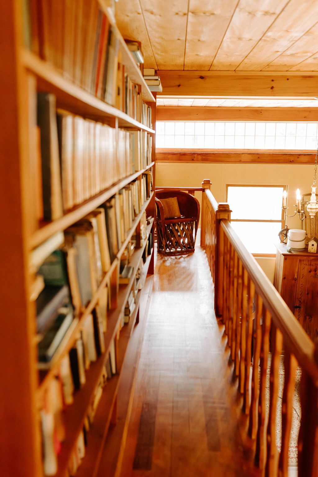 Wooden loft hallway with bookshelves, railing, and warm sunlight through windows