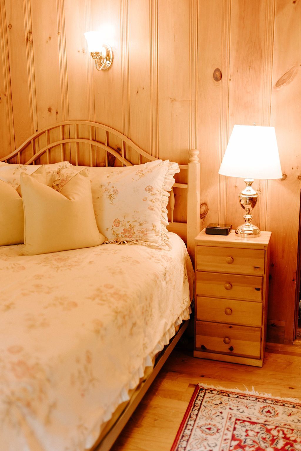 Cozy wood-paneled bedroom with floral bedding, bedside lamp, and small wooden nightstand