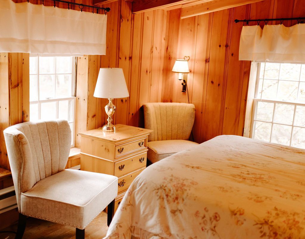 Cozy wood-paneled bedroom with two chairs, a lamp, and a floral bedspread by sunlit windows