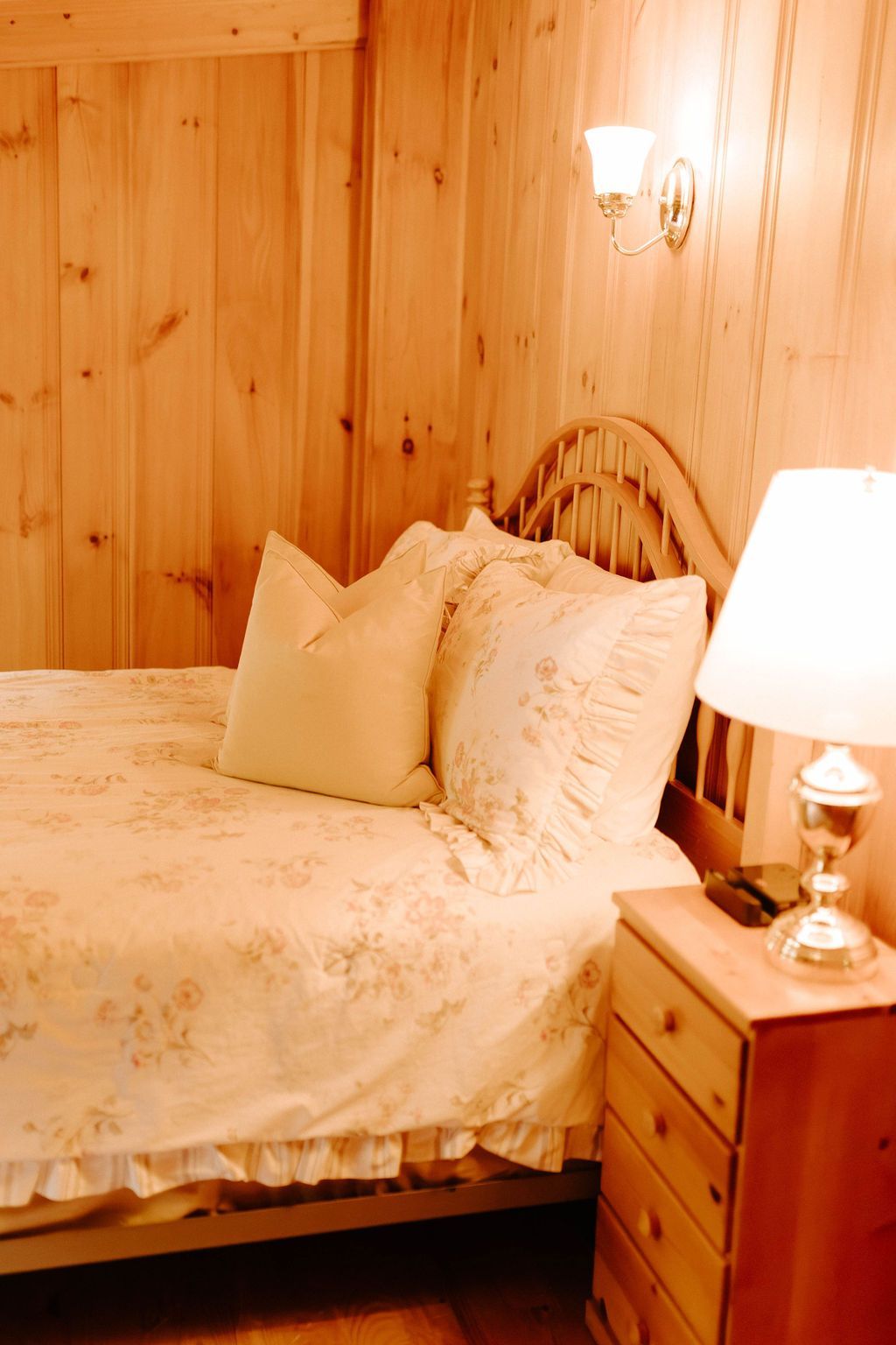 Cozy bedroom with a wooden bed, cream bedding, nightstand lamp, and wood-paneled walls.
