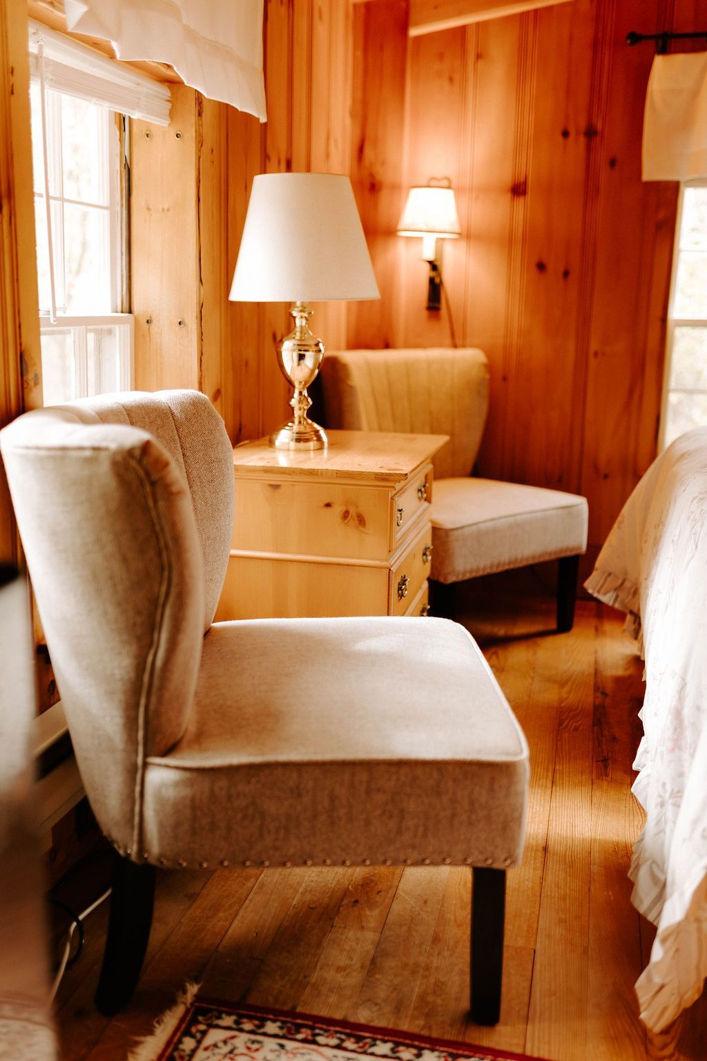 Cozy wood-paneled bedroom with upholstered chairs, bedside table, and lamps by a window
