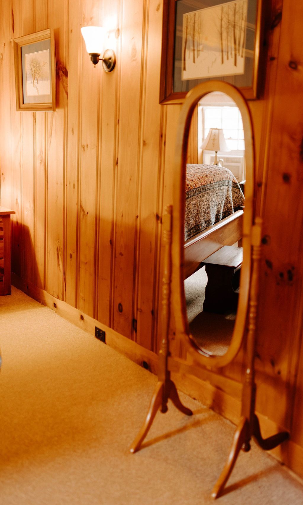 Wood-paneled room with a standing oval mirror reflecting a bed and lamp.