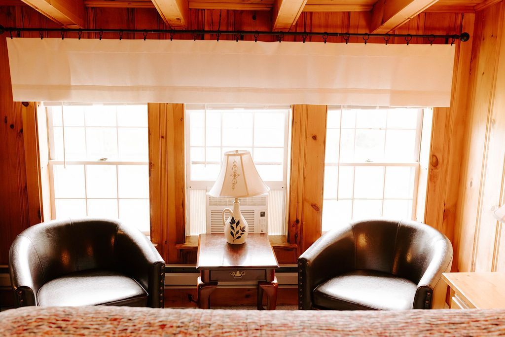 Cozy wood-paneled room with two black chairs, a lamp, and bright windows with curtains