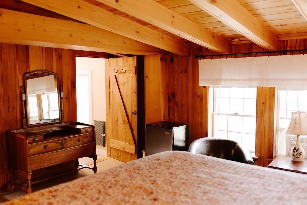Cozy wooden bedroom with exposed beams, dresser, mirror, window, and bed in the foreground