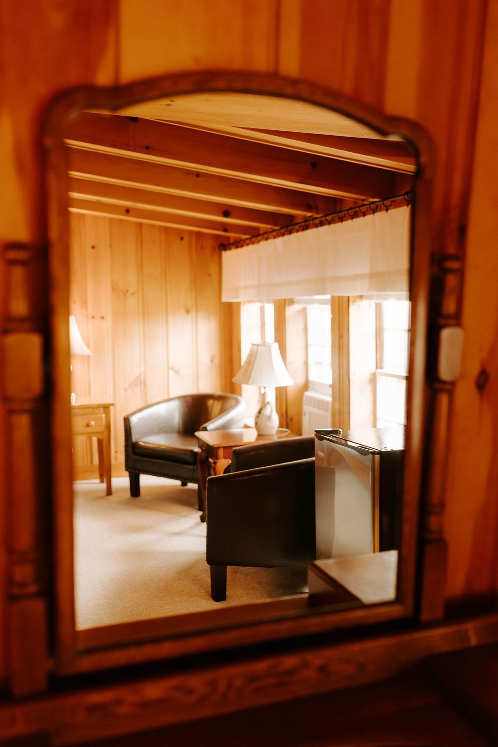 Mirror reflection of a cozy wood-paneled room with chairs, a lamp, and large windows
