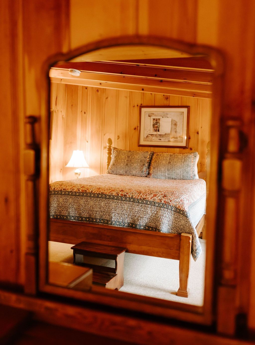 A cozy bedroom reflected in a framed mirror, with a lit bedside lamp and floral bedding.