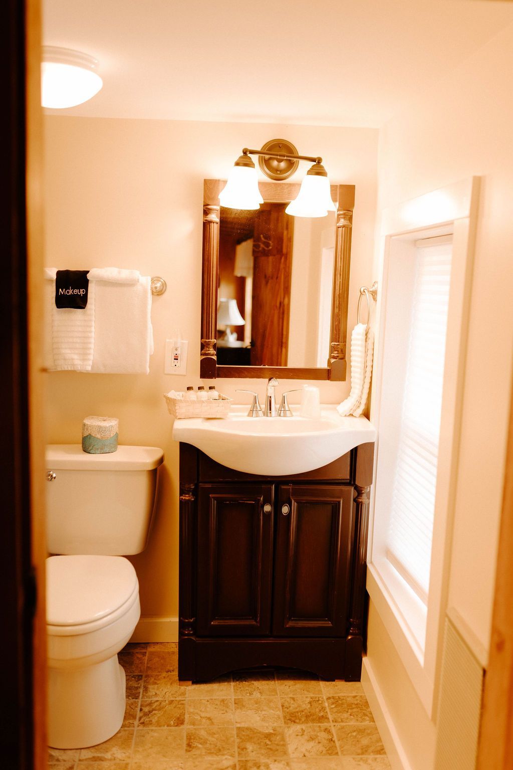 Narrow beige bathroom with toilet, sink vanity, mirror, and window blinds.