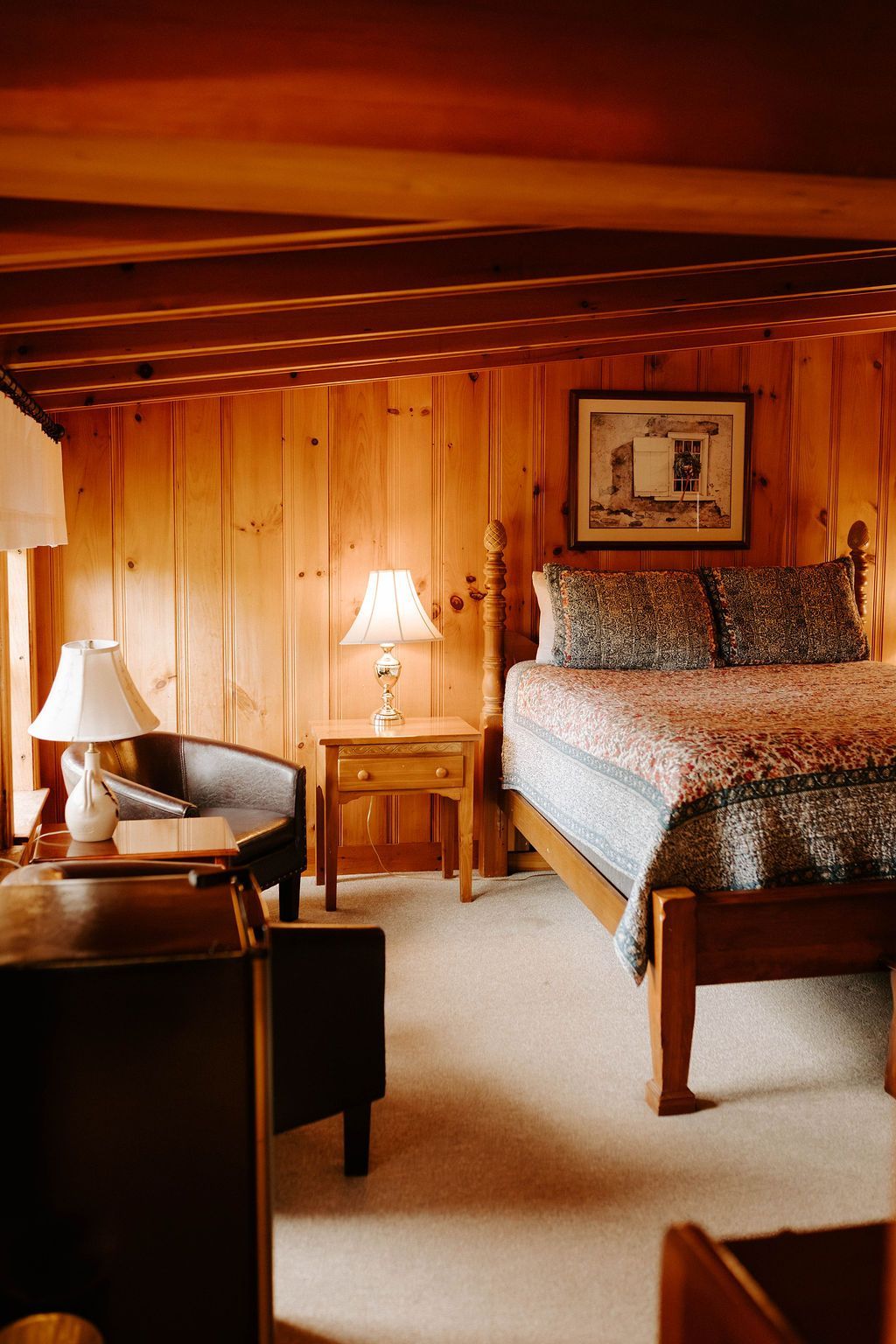 Cozy wood-paneled bedroom with a bed, bedside lamp, and armchair in warm orange light