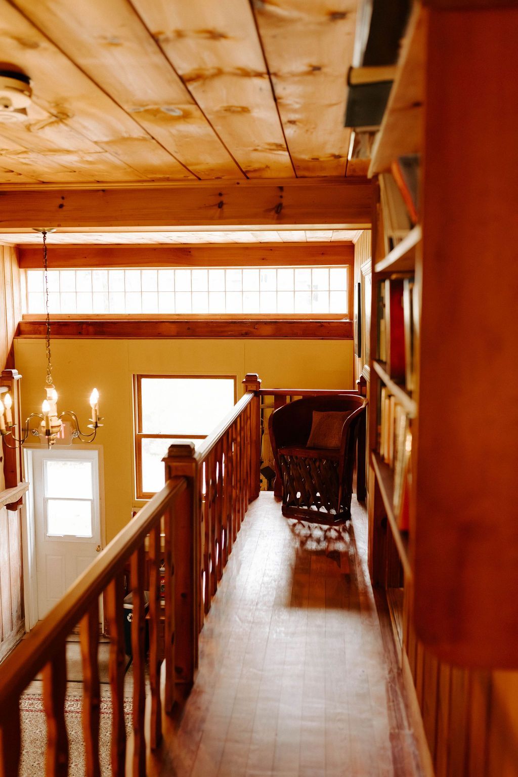 Wooden hallway with a staircase, balcony railing, and warm sunlight from windows.