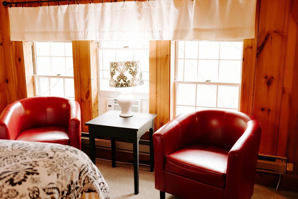 Cozy bedroom seating area with two red chairs, a bedside lamp, and bright windows with curtains