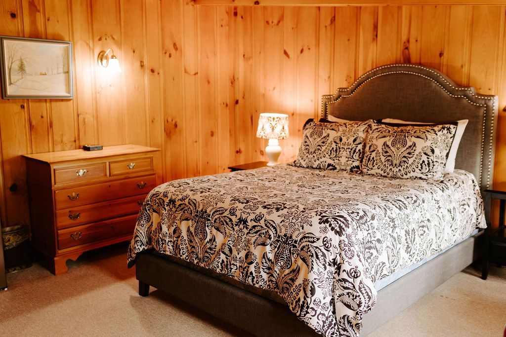 Cozy bedroom with wood-paneled walls, patterned bedspread, and matching dresser beside a bedside lamp.
