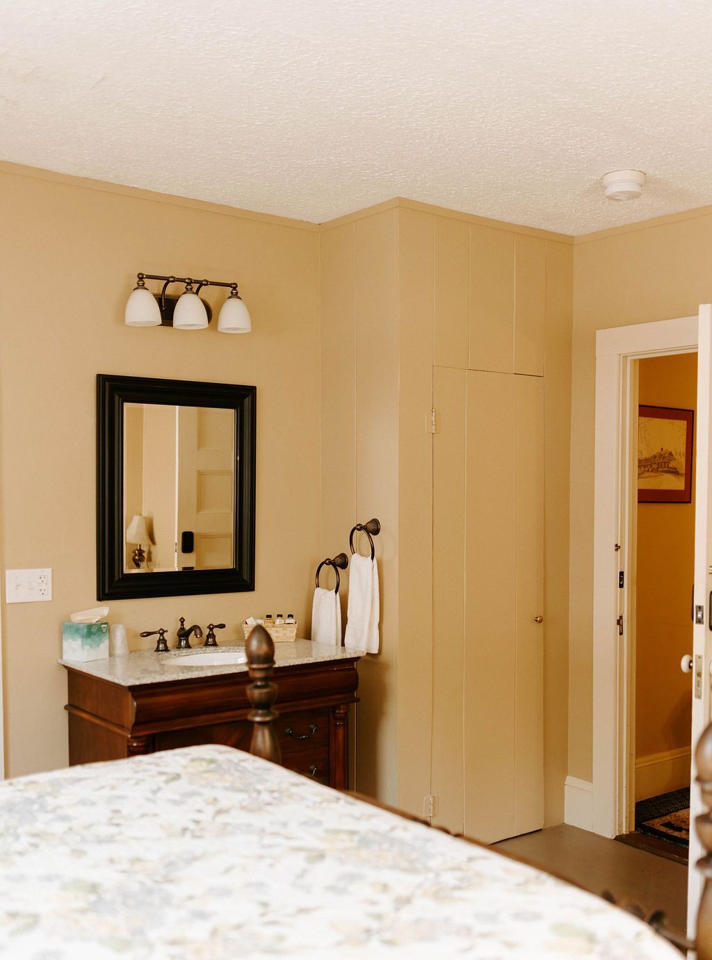 Bathroom vanity with mirror and light fixture in a beige room, seen from a bedroom doorway