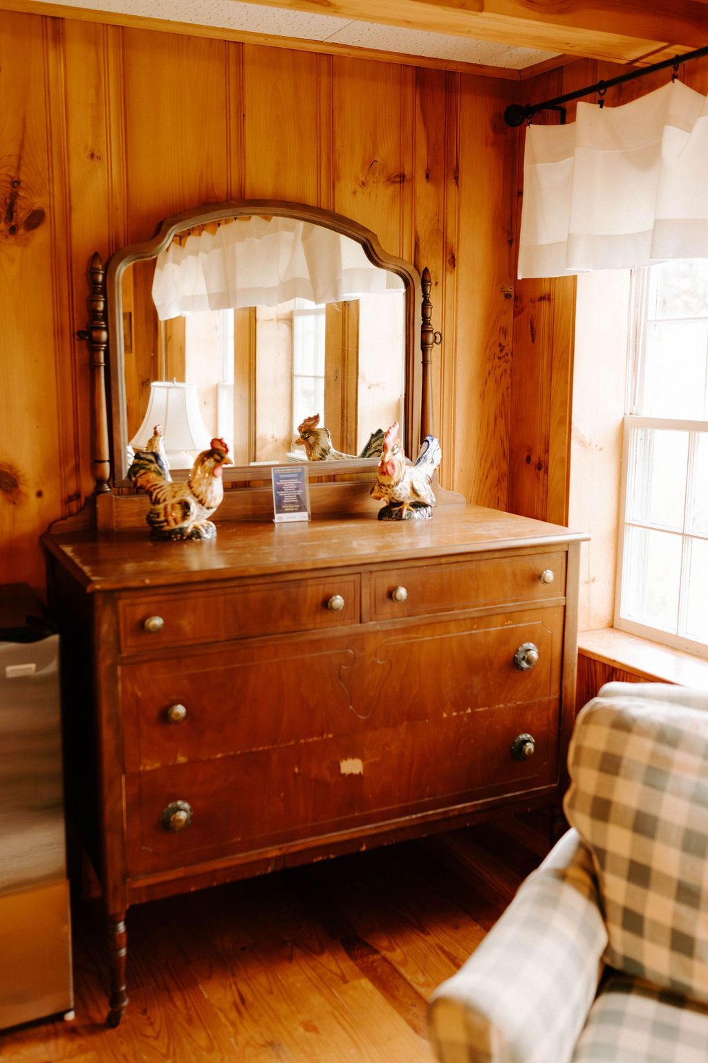 Wooden dresser with mirror in a sunlit room, beside a window and plaid chair