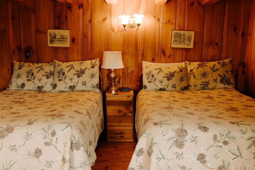 Two twin beds with floral quilts in a wood-paneled bedroom, separated by a nightstand and lamp.