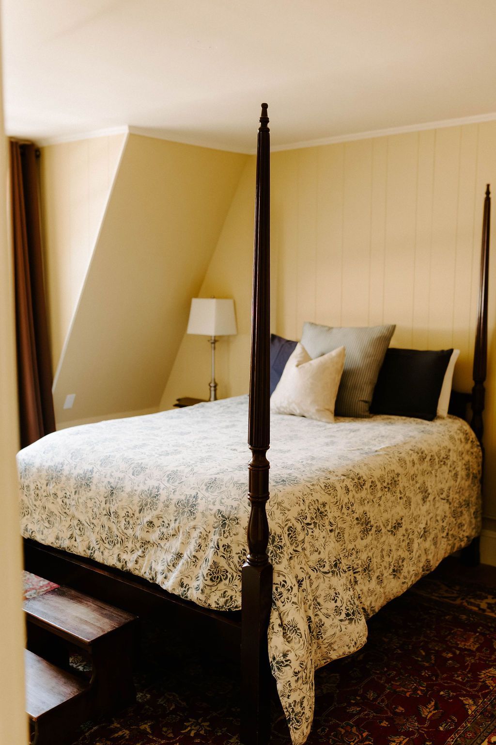 Four-poster bed with patterned quilt in a beige bedroom, beside a window and floor lamp.