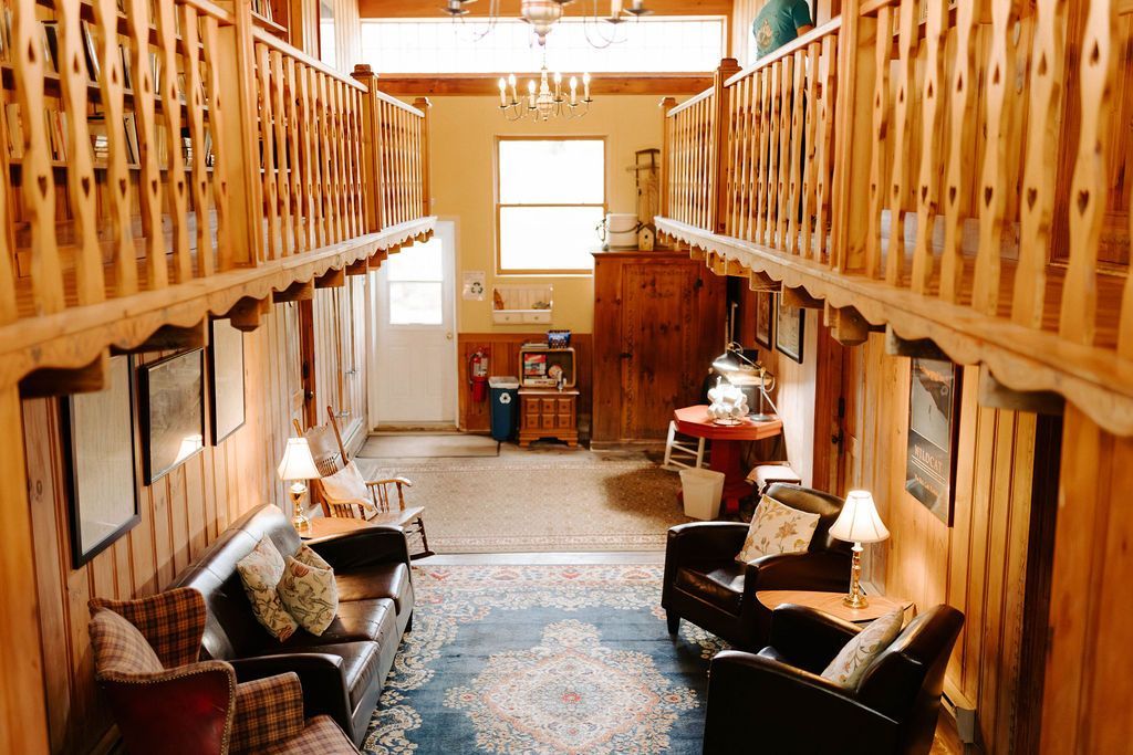 Wood-paneled lodge interior with loft railings, sofas, rug, and a bright window at the far end