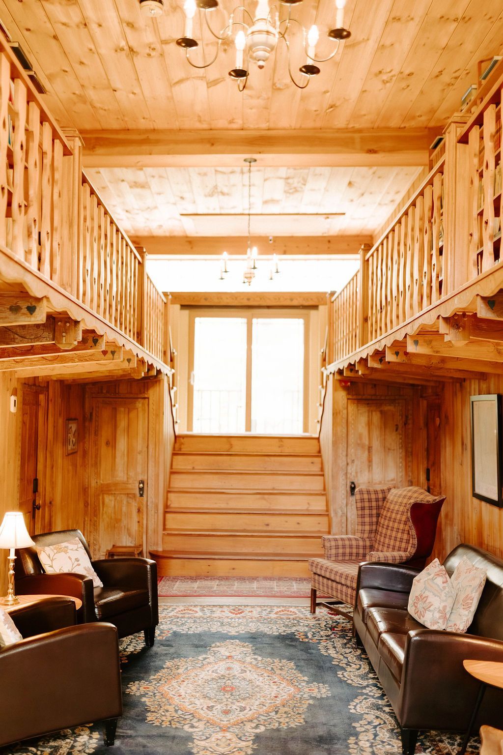 Rustic living room with wood-paneled walls, leather chairs, a patterned rug, and a staircase to an upper loft.