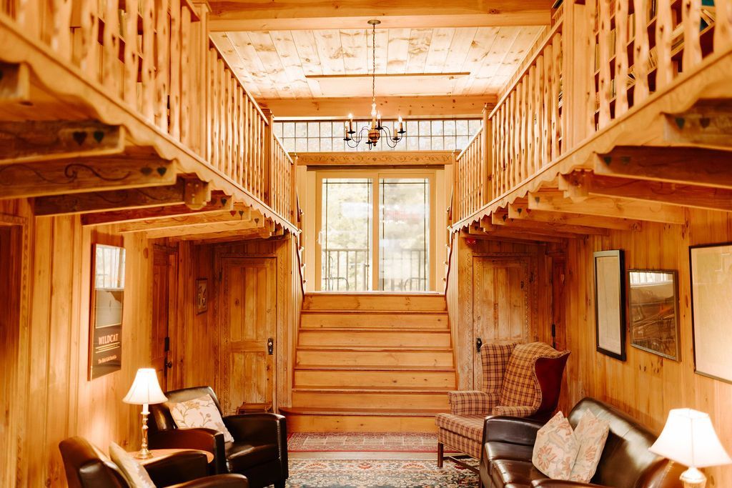 Wood-paneled lodge foyer with staircase, balcony, and warm lamps framing a central window