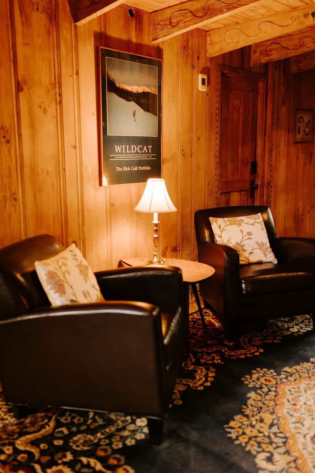 Cozy wood-paneled lounge with two black chairs, a small lamp table, and patterned rug.