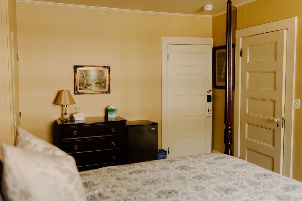Bedroom with beige walls, a bed in the foreground, and a dresser and framed picture against the back wall.