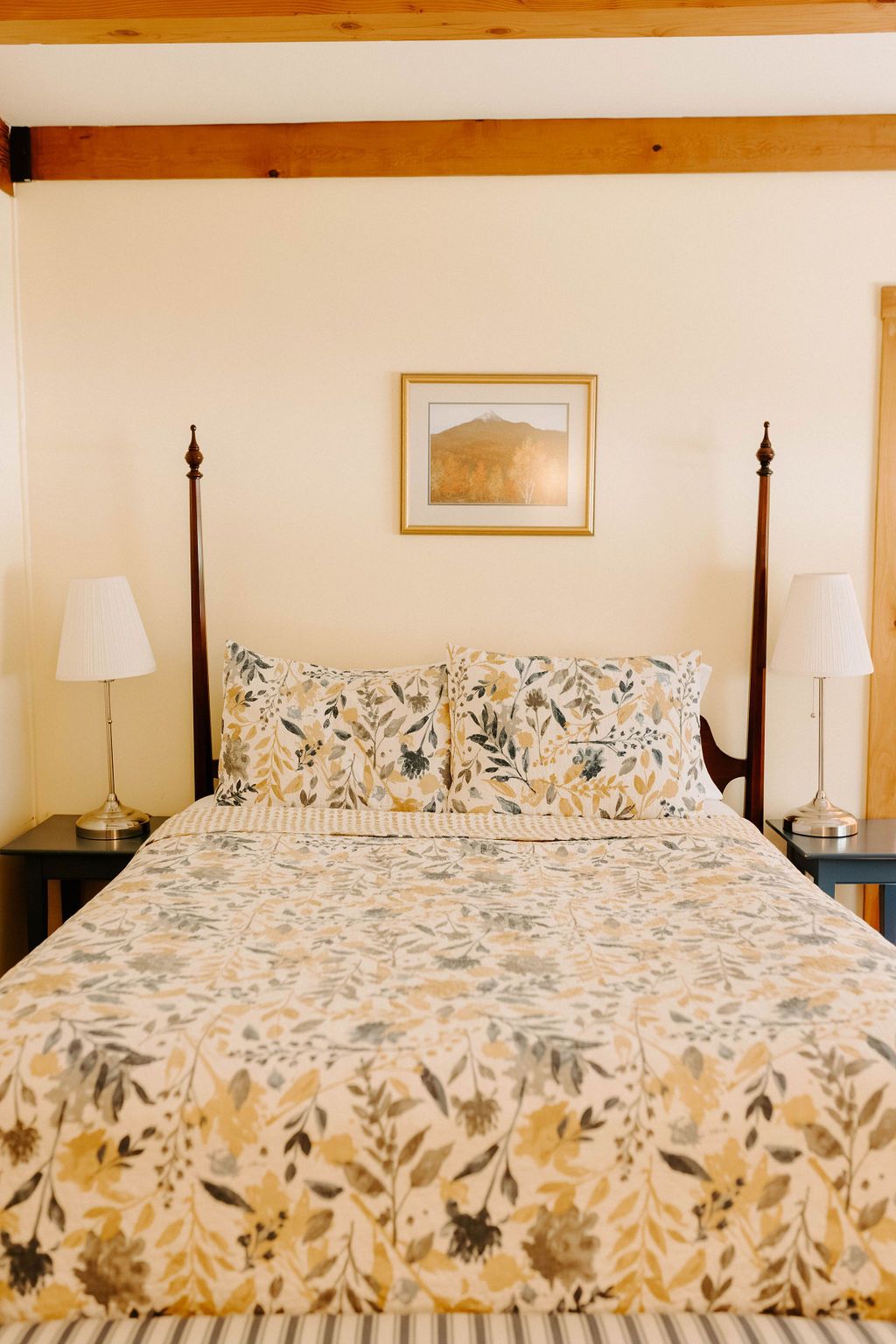 Floral-patterned bed with matching pillows in a cozy bedroom, flanked by lamps and dark wooden posts.