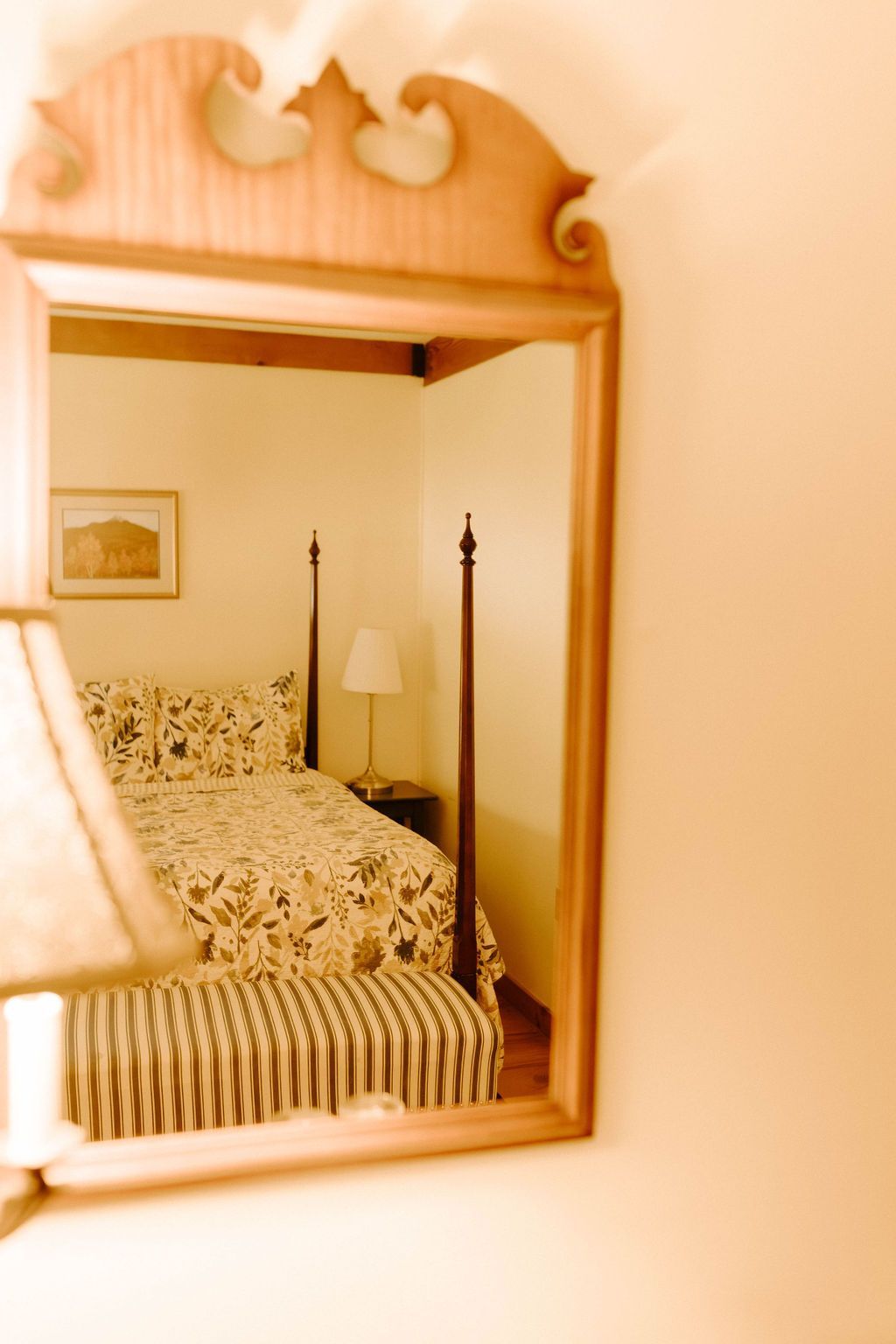 Bedroom reflected in a wooden-framed mirror, showing a bed with patterned bedding and a lamp beside it.