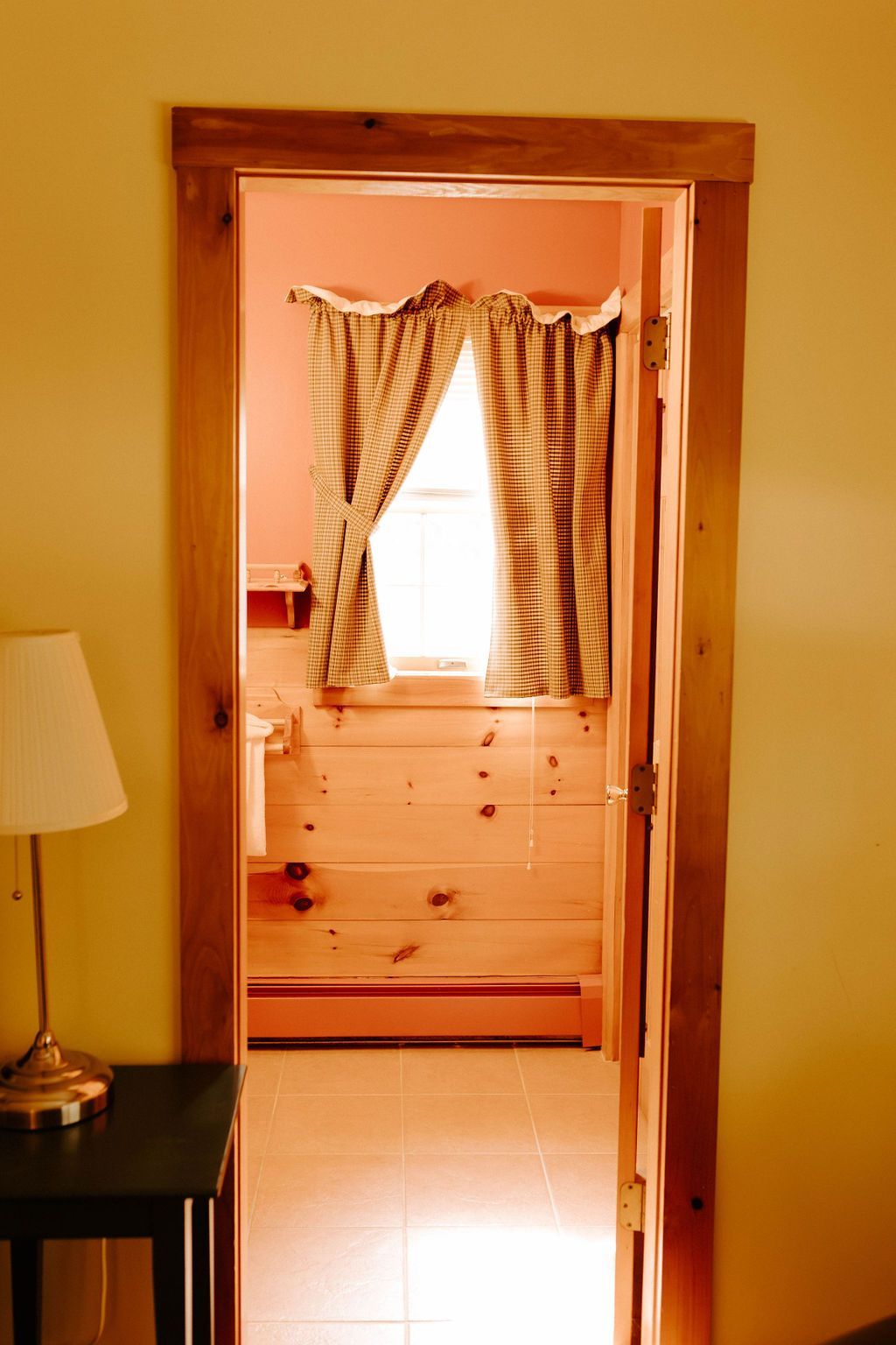 Wooden doorway leading to a sunlit room with plaid curtains and a dresser beneath the window