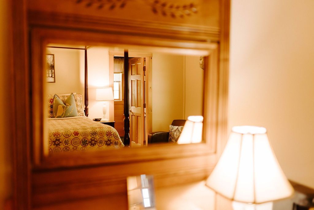 Warm bedroom reflected in a wooden-framed mirror, with a lit bedside lamp in the foreground.