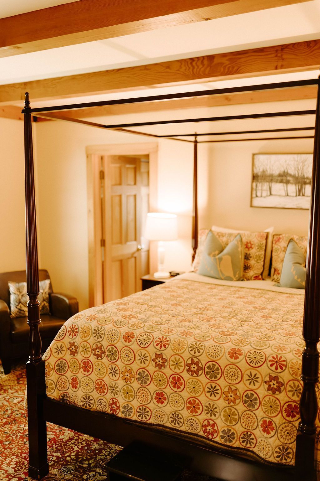 Cozy bedroom with four-poster bed, patterned quilt, bedside lamp, and wooden door in warm light