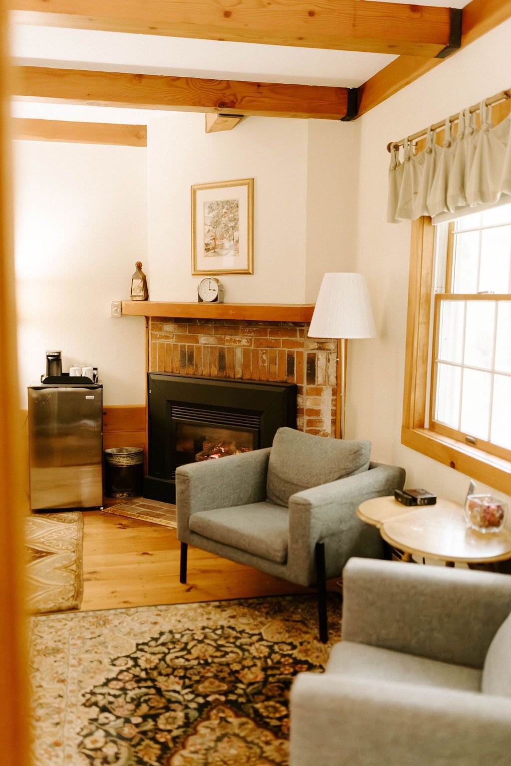 Cozy living room with exposed beams, fireplace, gray chairs, and sunlight through a window