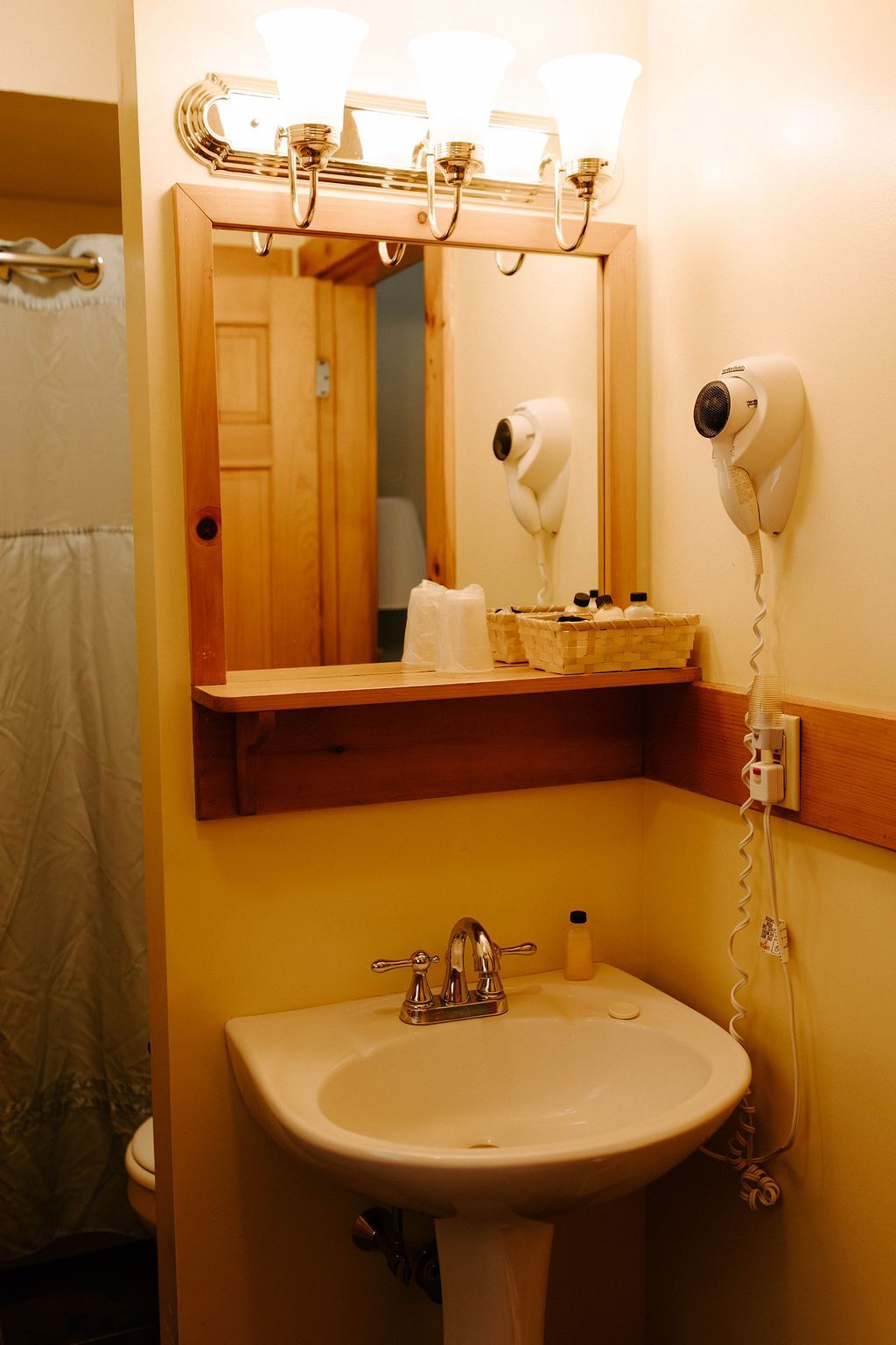 Small bathroom vanity with sink, mirror, lights, and wall-mounted hair dryers above a wood counter.