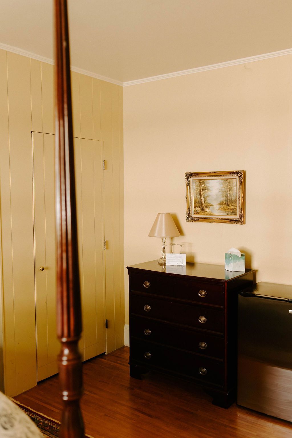 Bedroom corner with wooden dresser, lamp, mirror, and a bed beside a beige wall